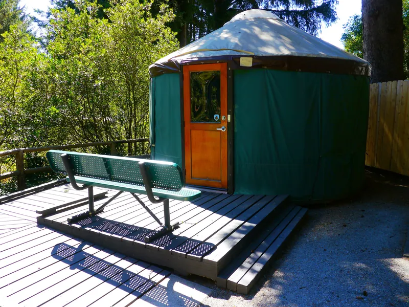 A green canvas yurt on a wooden deck with a bench, in the woods at a state park