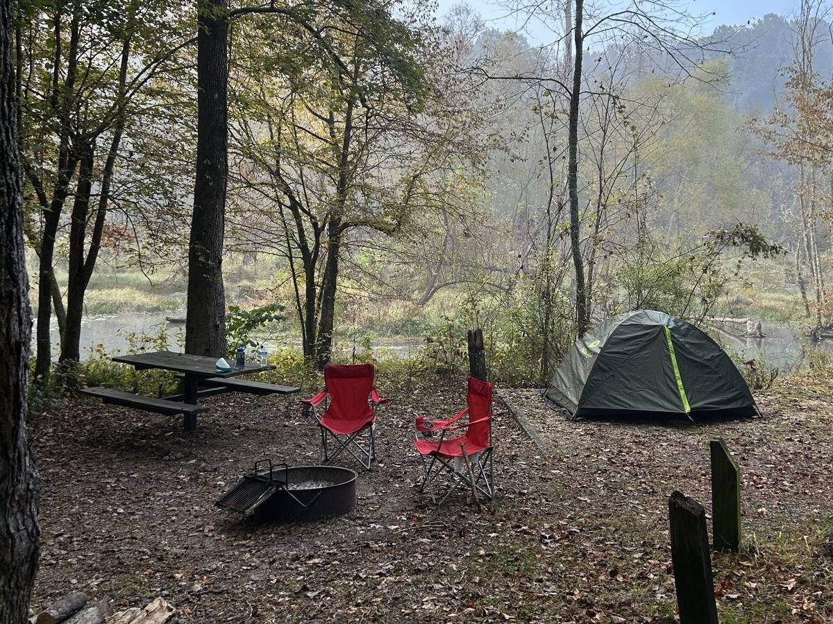 Tent, camp chairs, fire ring and picnic table at a Pocket Campground site with the misty beaver pond behind